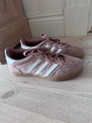 Adidas Gazelle rosa sneakers - Säljer ett par Adidas Gazelle sneakers i rosa mocka med vita detaljer och klassiska tre ränder på sidan. Skorna har gummisula och lågt skaft. Perfekta för dig som gillar retrostil och vill ha något som sticker ut lite extra.