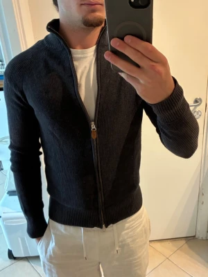 Mörkgrå stickad ziptröja från Morris  - Snygg mörkgrå stickad tröja med dragkedja framtill och hög krage. Tröjan har ribbade muddar vid ärmslut och nederkant, samt en stilren och enkel design som passar till det mesta. Perfekt för lager på lager och chill vardagsstil. Bra skick 