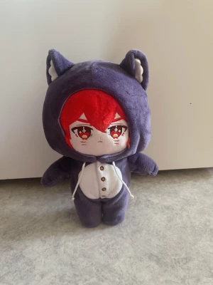 Söt Diluc kattdocka Dokidokicosplay - Mjukisdocka Diluc från Genshin Impact med rött hår och stora broderade ögon, klädd i en lila kattdräkt med öron, svans och vita detaljer. Dräkten har knappar framtill och snören vid huvan. Perfekt som dekoration eller samlarobjekt för animefans. Den är äkta från dokidokicosplay värde 600+ kr