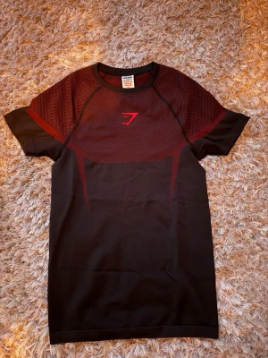 Svart och röd träningströja Gymshark - Snygg svart och röd träningströja från Gymshark med korta ärmar och rund hals. Tröjan har ett mesh-liknande mönster upptill och Gymshark-logga på bröstet. Tillverkad i ett stretchigt funktionsmaterial som andas, perfekt för gym eller löpning. 