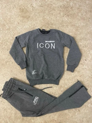 Grått loungewear-set ICON Ibrahimovic - Snyggt grått loungewear-set från Dsquared2 med 'IBRAHIMOVIC ICON' tryck på tröjan och byxan. Tröjan har en stor grafisk bild på ryggen och ribbade muddar. Byxorna har dragsko i midjan och dragkedjefickor. Perfekt för chill dagar hemma.