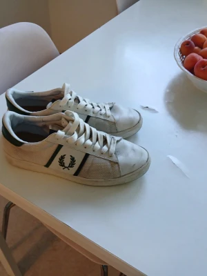 Vita Fred Perry sneakers med gröna detaljer - Snygga vita sneakers från Fred Perry med gröna och marinblå detaljer på sidan samt den klassiska lagerkransen. Skorna har lågt skaft, vita snören och ovandel i mesh och läder. Perfekta för en clean och sportig stil.