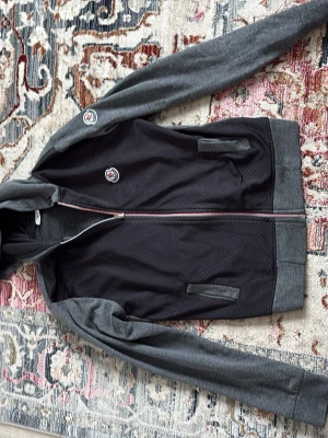 Moncler hoodie i svart och grå - Snygg hoodie från Moncler i svart med grå ärmar, huva och muddar. Dragkedja framtill och två fickor. Moncler-logga på bröstet och axeln. Materialet är en mix av bomull och polyester med meshstruktur på framsidan. Perfekt för en chill och sportig look. Handtaget på dragkedjan är lite sönder men den går fortfarande att dra upp och ner snabbt och smidigt.