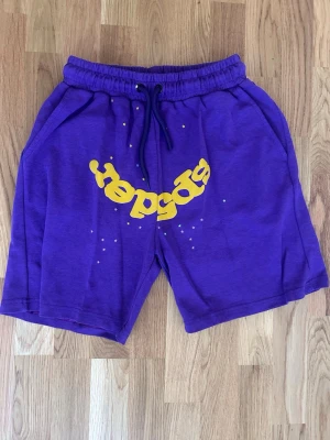 Sp5der Lila shorts - Säljer ett par lila shorts från Sp5der med gul tryckt logga framtill och små vita stjärndetaljer. Shortsen har resårmidja med snörning och är gjorda i mjuk bomull, perfekta för en avslappnad stil.