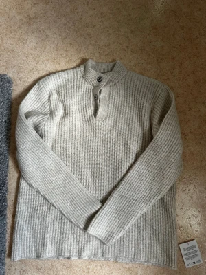 Tröja med Krage och Knappar - Stickad tröja i beige färg från Asos Design med ribbad struktur och hög krage. Säljs för den är i fel storlek för mig. Helt ny endast testad. Säljs för 299kr. Storlek M och sitter som M. Kontakta mig om du har frågor eller vill lägga bud.