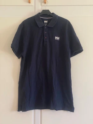 Mörkblå polo från Helly Hansen - Säljer en mörkblå polo från Helly Hansen med klassisk krage och knappar framtill. Skjortan har korta ärmar och logotyp på bröstet. Perfekt för dig som gillar sportig och enkel stil.