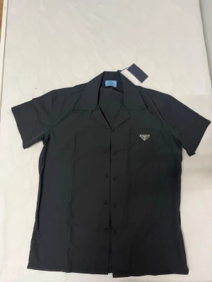 Svart kortärmad skjorta från Prada - Stilren svart skjorta från Prada med korta ärmar och klassisk krage. Skjortan har knappar framtill och en diskret logotyp på bröstet. Perfekt för dig som gillar minimalistisk design och exklusiva märken.