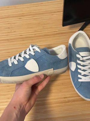 Blå Philippe Model sneakers i mocka - Snygga blå sneakers från Philippe Model i mjuk mocka med vita detaljer och vita skosnören. Skorna har en låg siluett, vit gummisula och ikonisk sköldlogga på sidan. Perfekta för dig som gillar stilrena och trendiga sneakers.