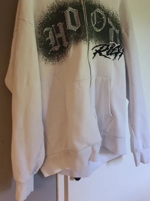 Vit hoodie med svart tryck och dragkedja - Vit hoodie med svart sprayliknande tryck och texten 'HOOD RICH' i gotisk stil över bröstet. Hoodien har dragkedja framtill, känguruficka och huva. Materialet ser ut att vara mjuk bomullsblandning, perfekt för en streetwear-look. Kan sänka priset vid snabb affär!