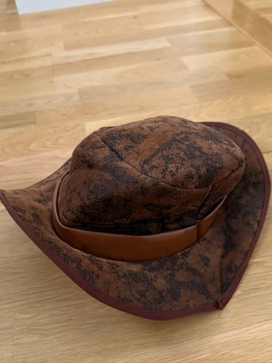 Brun cowboyhatt med läderband - Cool cowboyhatt i brun färg med mörka mönster och ett läderband runt kronan. Hatten har breda brätten och är tillverkad i ett mjukt tygmaterial som ger en avslappnad look. Perfekt för dig som vill sticka ut med en westernstil.