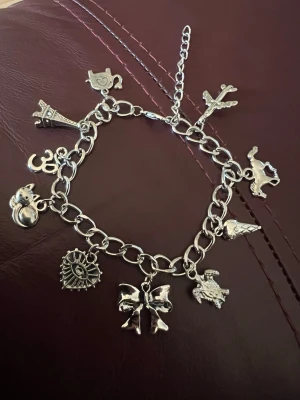 Silverfärgat berlockarmband med charms - Charmigt armband i silverfärgad metall med flera olika berlocker, bland annat katt, rosett, hjärta, flygplan, tekanna och Eiffeltorn. Länkarna är grova och armbandet har justerbar kedja. Perfekt för dig som gillar unika detaljer och personliga smycken.
