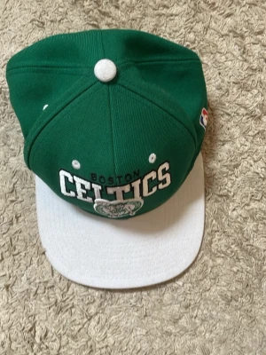 Grön Boston Celtics keps - Säljer en grön och vit Boston Celtics keps med broderad logga och text framtill. Kepsen har platt skärm i vitt och klassisk snapback-modell. Materialet är bomull och kepsen har officiell NBA-märkning på sidan.