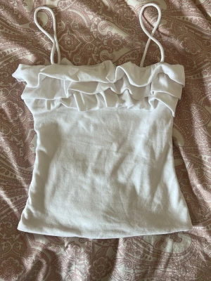 Vit volang topp från Zara - Söt vit linnetopp från Zara med tunna axelband och dubbla volangdetaljer upptill. Perfekt för varma dagar och enkel att styla med jeans eller kjol. Tillverkad i mjuk bomull för en bekväm känsla.