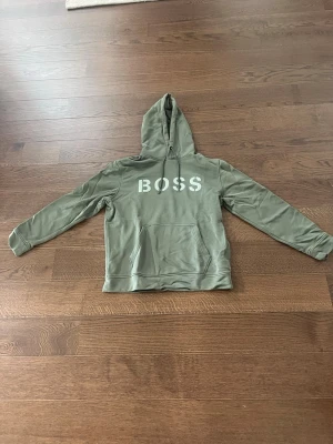 Grön hoodie från Hugo Boss - Snygg grön hoodie från Hugo Boss med stor vit BOSS-logga på bröstet. Hoodien har huva med dragsko och en stor magficka framtill. Tillverkad i mjukt bomullsmaterial som känns skönt mot huden. Perfekt för en avslappnad och trendig look.