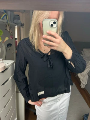 Svart blus med knyt från Odd Molly - Snygg svart blus från Odd Molly med knytband vid halsen och diskreta volangdetaljer vid ärmslut och nederkant. Tillverkad i ett tunt, lätt material som ger en luftig känsla. 