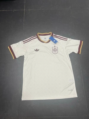 Vit Spanien tröja från Adidas - Säljer en vit Spanien fotbollströja från Adidas med vinröda och gula detaljer. Tröjan har korta ärmar, rund halsringning och tre klassiska Adidas-ränder på axlarna. På bröstet finns Spaniens landslagsemblem och Adidas-logga. Materialet är lätt och andas.