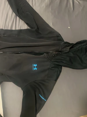 Svart hoodie från Under Armour - Svart hoodie från Under Armour med dragkedja och blå logga på bröstet. Tillverkad i mjukt och värmande material, perfekt för träning eller chill. Huva med dragsko och fickor framtill. Snygg och sportig look med diskreta detaljer. Dock en liten hål vid armen 