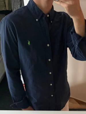 Mörkblå skjorta från Ralph Lauren - Klassisk mörkblå skjorta från Ralph Lauren med button-down krage och grönt broderat logotyp på bröstet. Skjortan har långa ärmar och vita knappar framtill. Perfekt för en clean och stilren look. Ganska urtvättad därav priset, annars bra skick 