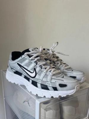 Nike vita och svarta sneakers - Nike sneakers i vitt och svart med mesh och syntetiska paneler. Skorna har en chunky sula, svart swoosh på sidan och vita skosnören. Sportig design med retrovibbar och detaljerade sömmar. Perfekta för dig som gillar streetwear och bekväma sneakers.