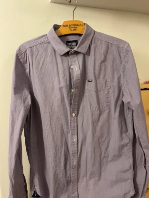 Ljuslila skjorta från Superdry - Klassisk ljuslila skjorta från Superdry med bröstficka och knappar framtill. Skjortan har lång ärm och en liten logotyp på bröstfickan samt på ärmen. Perfekt till jeans eller chinos för en clean look.