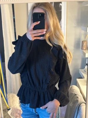 Svart volangblus från Lindex - Trendig svart blus från Lindex med volangdetaljer framtill och vid axlarna. Blusen har en lätt skrynklig struktur, markerad midja och långa ärmar. Perfekt att styla med jeans eller kjol för en cool look.skulle säga att den passar en xxs/xs