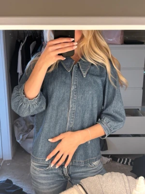 Klassisk blå jeansjacka med krage - Snygg blå jeansjacka med markerad krage och trekvartsärmar med resår vid ärmslut. Jackan har en rak passform och stängs framtill med dragkedja som är dold. Perfekt att styla till jeans för en cool denim-on-denim look.