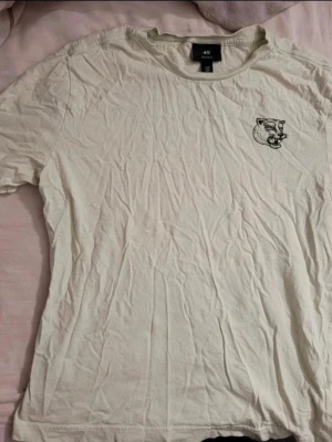 T-shirt med tryck från H&M - Ljusgrön t-shirt från H&M med ett litet svart tryck av ett djurhuvud på bröstet. Klassisk passform och rund halsringning. Perfekt basplagg för alla tillfällen.
