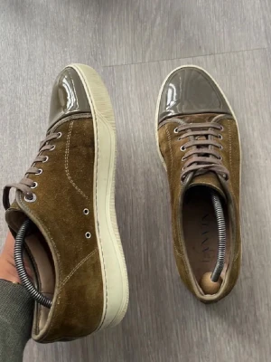 Bruna sneakers med lackad tå - Snygga sneakers från Lanvin i brunt mocka med glansig, lackad tå i grått. Skorna har grå snörning och vit sula. Perfekt för dig som gillar en unik och trendig stil med mix av material och färger.