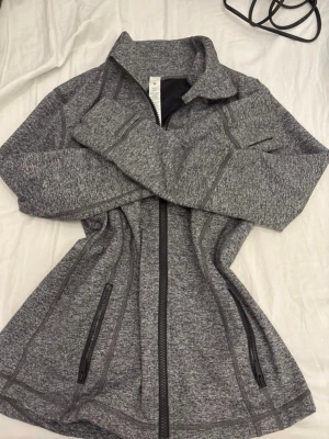 Lululemon zip up - Snygg gråmelerad sportjacka med hel dragkedja framtill och två fickor med dragkedja. Jackan har hög krage och figurnära passform, perfekt för träning eller promenader. Tillverkad i mjukt och stretchigt material för maximal komfort.