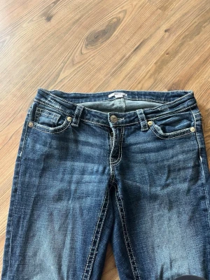 Blå jeans med broderade bakfickor - Säljer ett par blå jeans med snygg slitning och kontrastsömmar. Jeansen har fem fickor och coola broderier på bakfickorna som ger extra stil. Passar perfekt till en avslappnad look och har klassisk knappgylf och bälteshällor.