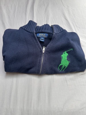 Marinblå ziptröja Polo Ralph Lauren - Marinblå stickad ziptröja från Polo Ralph Lauren med stor grön logga på bröstet. Tröjan har hög krage och grovstickad struktur, perfekt för en avslappnad men ändå snygg look. Klassisk design med dragkedja framtill. För tjejer: Sitter oftast perfekt som en XS eller mindre S. Det är superpopulärt bland tjejer att köpa Ralph Laurens barn-/ungdomsstorlekar eftersom de ger en lite mer slimmad och croppad passform än herrmodellerna. För killar/ungdomar: Passar bäst för killar i 14–16 årsåldern (ca 158
