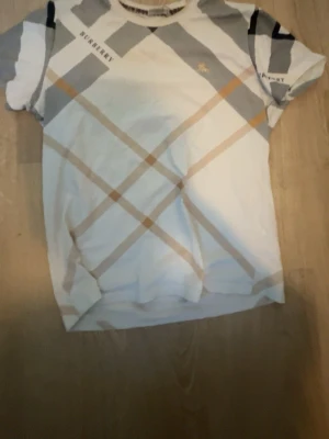 Vit rutig t-shirt från Burberry - Snygg vit t-shirt från Burberry med klassiskt rutigt mönster i beige och grått. T-shirten har korta ärmar och rund halsringning. Burberry-logga broderad på bröstet och tryckt på axeln. Perfekt för dig som gillar stilrena designer med lyxig känsla.
