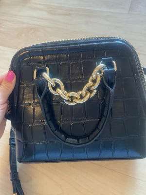 Svart handväska med kedjedetalj - Snygg svart handväska i croco-mönstrat konstläder med guldfärgad kedja som detalj på handtaget. Väskan har en fyrkantig form och dragkedja upptill. 