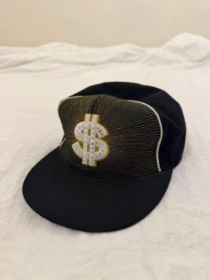 Klassisk New Era svart keps, storlek 7 1/4 - Svart vintage New Era keps med en guldfärgad dollarteckenbrodyr på framsidan, prydd med strassdetaljer. Samma dollartecken finns även broderat på sidan av kepsen. Kepsen har en platt skärm och är designad med en 59FIFTY-passform.