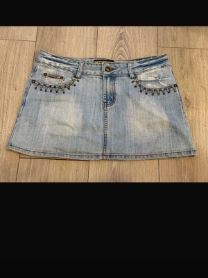 Ljusblå jeanskjol med nitar - Trendig ljusblå jeanskjol från Replay med dekorativa nitar vid fickorna framtill och broderade detaljer bak. Klassisk femficksmodell i kort längd och lätt tvättad denim. Perfekt för dig som gillar en cool och avslappnad stil.