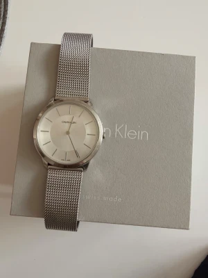 Silverfärgad klocka från Calvin Klein - Stilren klocka från Calvin Klein med rund urtavla i vitt och minimalistiska markeringar. Armbandet är i mesh och har en snygg silverfinish som ger en modern look. Perfekt accessoar för dig som gillar enkel och tidlös design. Batteriet på klockan behöver bytas!