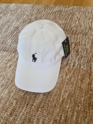 Vit keps från Polo Ralph Lauren - Snygg vit keps från Polo Ralph Lauren med klassisk broderad logga framtill och justerbart spänne bak. Kepsen är i bomull och har en böjd skärm. Perfekt accessoar för en clean och sportig look.
