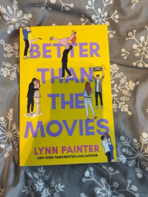 Better Than the Movies bok - En färgstark pocketbok med gul framsida och lila text, dekorerad med tecknade figurer i romantiska scener. Boken är skriven av Lynn Painter och är en romantisk komedi som handlar om tonårskärlek och drömmar om det perfekta filmögonblicket.