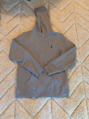 Grå hoodie från Polo Ralph Lauren - Snygg grå hoodie från Polo Ralph Lauren med klassisk broderad logga på bröstet. Hoodien har huva, känguruficka och är tillverkad i mjukt bomullsmaterial. Perfekt för en chill och avslappnad stil. Skulle säga att den passar dig som är mellan 170-180 beroende på vikt