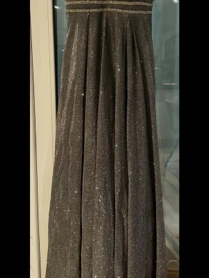 Glittrig långklänning i silver - Säljer en elegant långklänning i glittrigt silvertyg. Klänningen har ett svepande fall och är perfekt för dig som vill sticka ut med en glamorös look. Materialet är mjukt och skimrande, vilket ger en lyxig känsla.