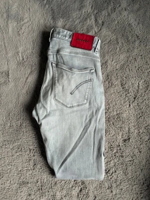 grå Dondup jeans - Säljer ett par ljusgrå jeans från Dondup med klassisk femficksdesign och en snygg röd patch med logga bak i midjan. Det är modell George och sitter som ”slim fit”. Mycket bra skick knappt använda. 
