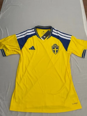 Svensk landslagströja Adidas gul/blå - Gul och blå fotbollströja från Adidas med Sveriges landslagsemblem på bröstet. Tröjan har korta ärmar, blå detaljer på axlarna och vita ränder. Perfekt för fotboll och träning.