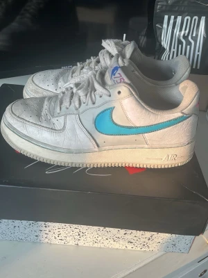 Nike Air Force 1 med blå swoosh - Säljer ett par klassiska Nike Air Force 1 sneakers i vitt läder med en blå swoosh på sidan. Skorna har gråa skosnören, perforerad tåbox och en vit sula med AIR-detalj. På plösen finns en 75-års logga. Perfekta för dig som gillar retrostil och streetwear.