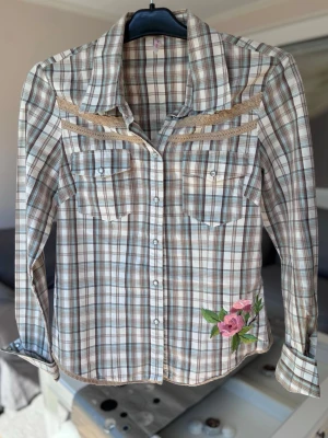Rutig skjorta med broderad blomma - Snygg rutig skjorta i beige, brun och blå toner med långa ärmar och två bröstfickor. Skjortan har dekorativa spetsdetaljer och en broderad rosa blomma nertill. 