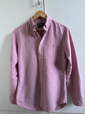 Rosa skjorta från Polo Ralph Lauren - Klassisk rosa skjorta från Polo Ralph Lauren med button-down krage och broderad logga på bröstet. Skjortan har långa ärmar, knappar framtill och är tillverkad i mjuk bomull. Perfekt för en sommardags! 