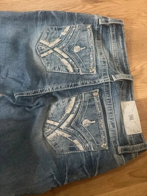 Miss Me blå jeans med detaljer - Säljer ett par blå Miss Me jeans med slitningar och snygga bakfickor dekorerade med paljetter och broderier. Jeansen har silverfärgade knappar och kontrastsömmar som ger en cool look. Perfekta för dig som gillar statement-jeans!