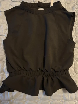 Svart ärmlös topp med volang - Snygg svart ärmlös topp med hög krage och volang nedtill. Toppen har resår i midjan för en markerad siluett och knappar i ryggen som detalj. Perfekt för dig som vill ha en stilren men ändå unik look.