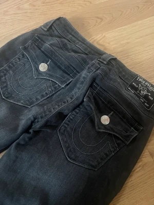 Svarta True Religion jeans - Säljer ett par svarta jeans från True Religion, modell Becca. Jeansen har stora bakfickor med ikoniska vita sömmar och silverfärgade knappar. Materialet är mjukt denim och passformen är slim med låg midja. Perfekta för dig som gillar streetwear och snygga detaljer.