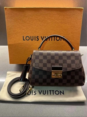 Louis Vuitton Damier handväska brun - Säljer en snygg handväska från Louis Vuitton i klassiskt brunt Damier-mönster. Väskan har guldfärgade metalldetaljer, ett topphandtag och avtagbar axelrem. Tillverkad i canvas med läderdetaljer och elegant lås framtill. Perfekt för dig som gillar lyxig design.