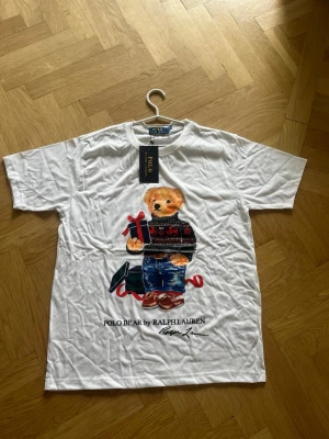 Vit Polo Bear t-shirt från Ralph Lauren - Vit t-shirt från Ralph Lauren med ikoniskt Polo Bear-tryck framtill. Björnen har en julig tröja och håller i ett paket. Klassisk passform och rund hals. Perfekt för dig som gillar statement-plagg och vill sticka ut lite extra. Jag är 181cm och den sitter perfekt på mig👍 jag skickar samma dag, kontakta för minsta lilla fundering! (100% cotton)